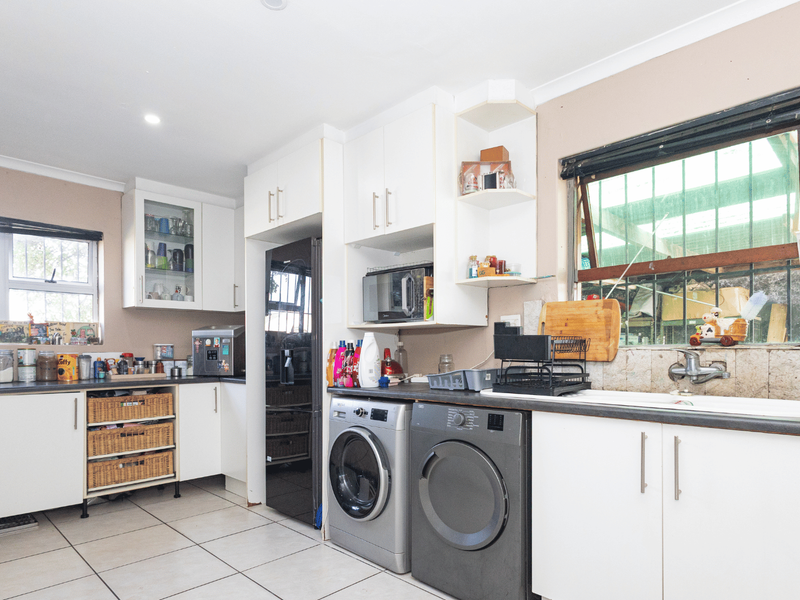 A functional kitchen featuring white cabinetry, laminate countertops, a double sink, and integrated laundry appliances, with tiled flooring and security bars on the windows.