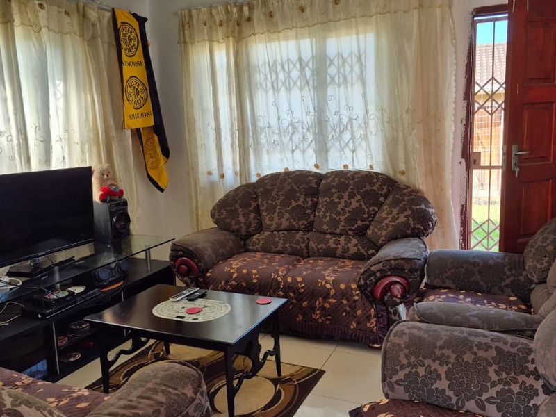 A living room featuring tiled flooring, a timber entrance door with a security gate, and large windows with curtains.