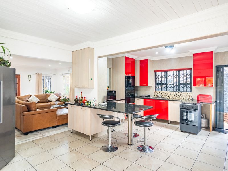 Open-plan kitchen and living area featuring high-gloss red and light wood cabinetry, granite countertops, a breakfast bar, and tiled flooring throughout.