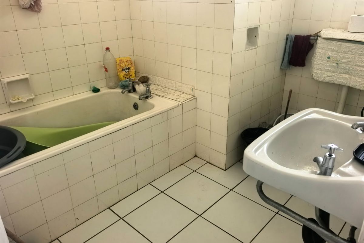 A dated bathroom featuring a built-in bathtub, pedestal washbasin, and full-height ceramic wall tiling, requiring modernization of finishes and fixtures.