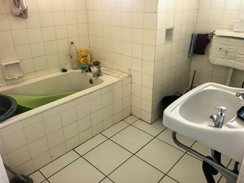 A dated bathroom featuring a built-in bathtub, pedestal washbasin, and full-height ceramic wall tiling, requiring modernization of finishes and fixtures.