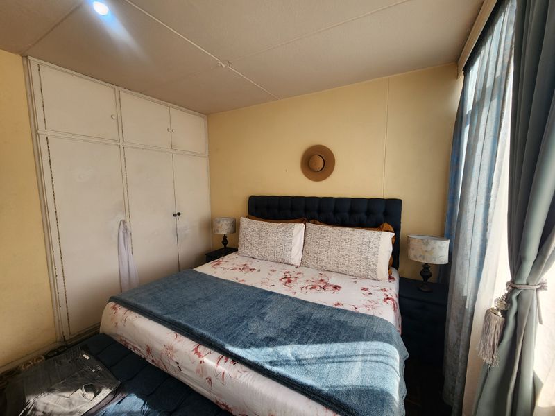 A bedroom featuring built-in cupboards and large windows. The room shows signs of wear, including visible ceiling cracks and dated finishes.