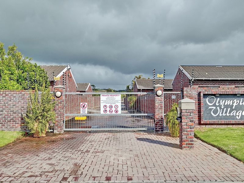 Secure gated entrance to Olympic Village townhouse complex, featuring brick perimeter walls, electric fencing, and a paved driveway.