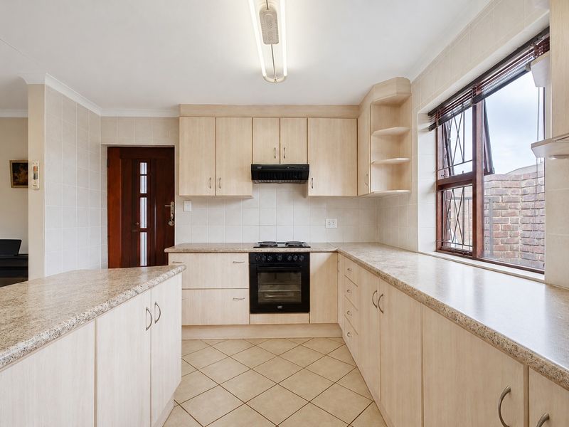 A functional kitchen featuring light wood-finish cabinetry, laminate countertops, built-in oven and hob, tiled flooring, and a window with security bars.