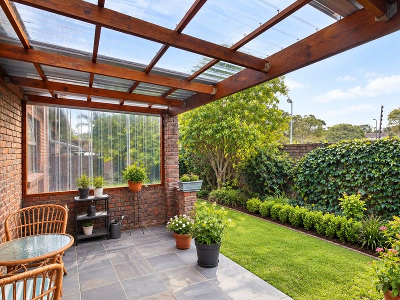 A well-maintained covered patio featuring a timber pergola with polycarbonate roofing, tiled floors, and direct access to a landscaped garden with boundary walls.