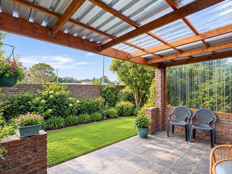 A well-maintained covered patio featuring a timber pergola with corrugated roofing, brick pillars, and tiled flooring, overlooking a landscaped garden with a brick boundary wall.
