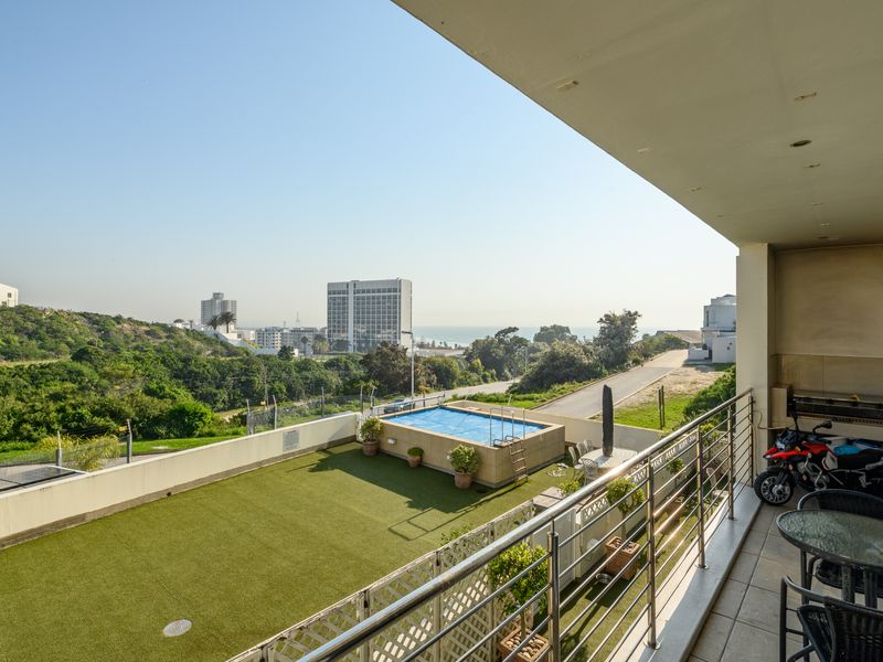 Spacious covered balcony featuring stainless steel balustrades and tiled flooring, overlooking a private garden area with a raised swimming pool and coastal views.