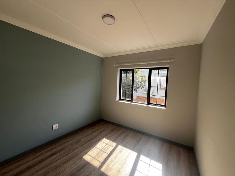 A bright, empty bedroom featuring modern laminate flooring, a neutral color palette with an accent wall, and a window with security bars.