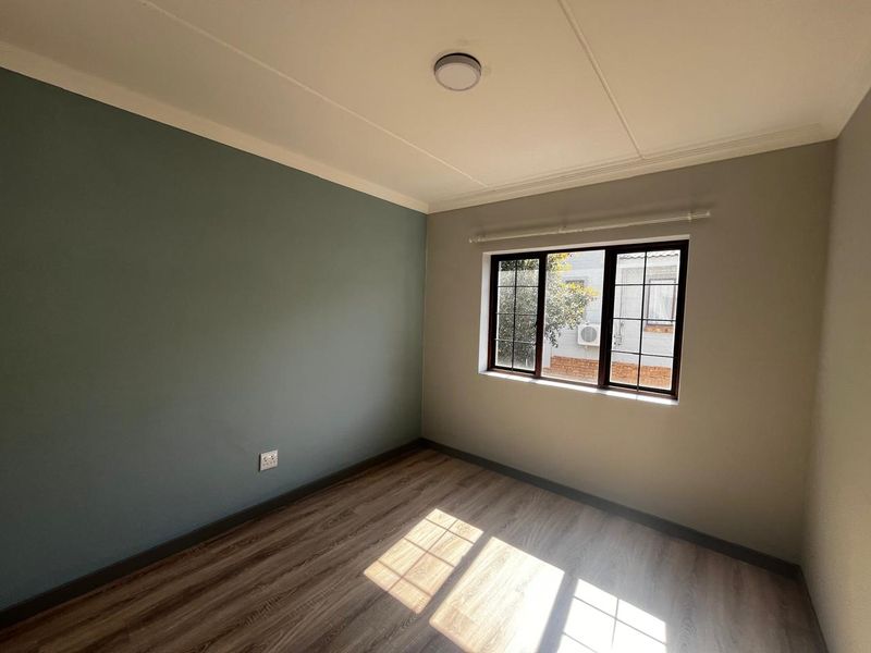 A bright, empty bedroom featuring modern laminate flooring, a dual-tone painted wall, and a window fitted with security bars.