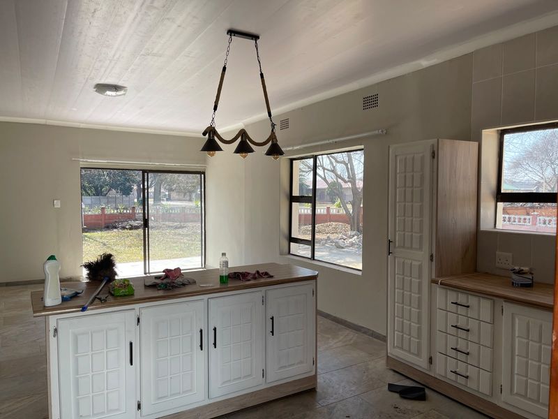 A bright, open-plan kitchen featuring a central island, ample built-in cabinetry, tiled flooring, and large windows providing natural light and garden views.