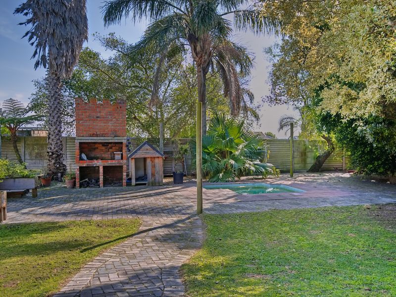 Rear garden featuring a paved patio area, a built-in brick braai, and a swimming pool surrounded by mature trees and boundary walling.