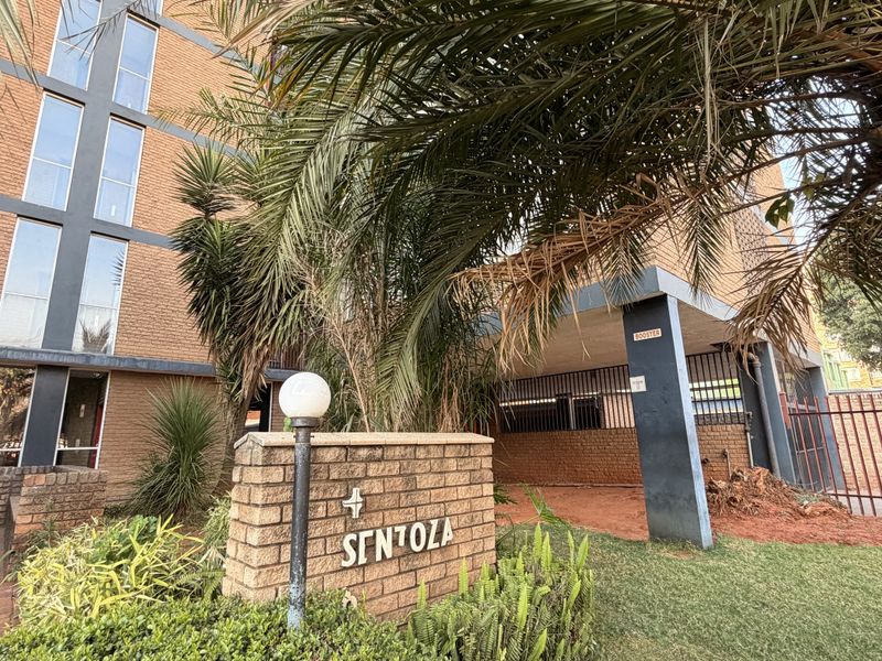 Front entrance view of the Sentola apartment complex, featuring a brick facade, paved driveway, and established garden landscaping.
