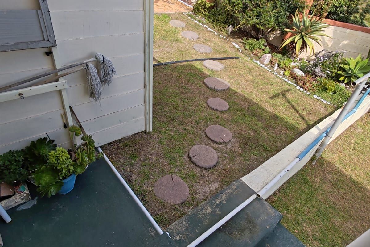 A rear garden area featuring concrete steps leading down to a lawn with a stepping stone path and perimeter boundary wall.