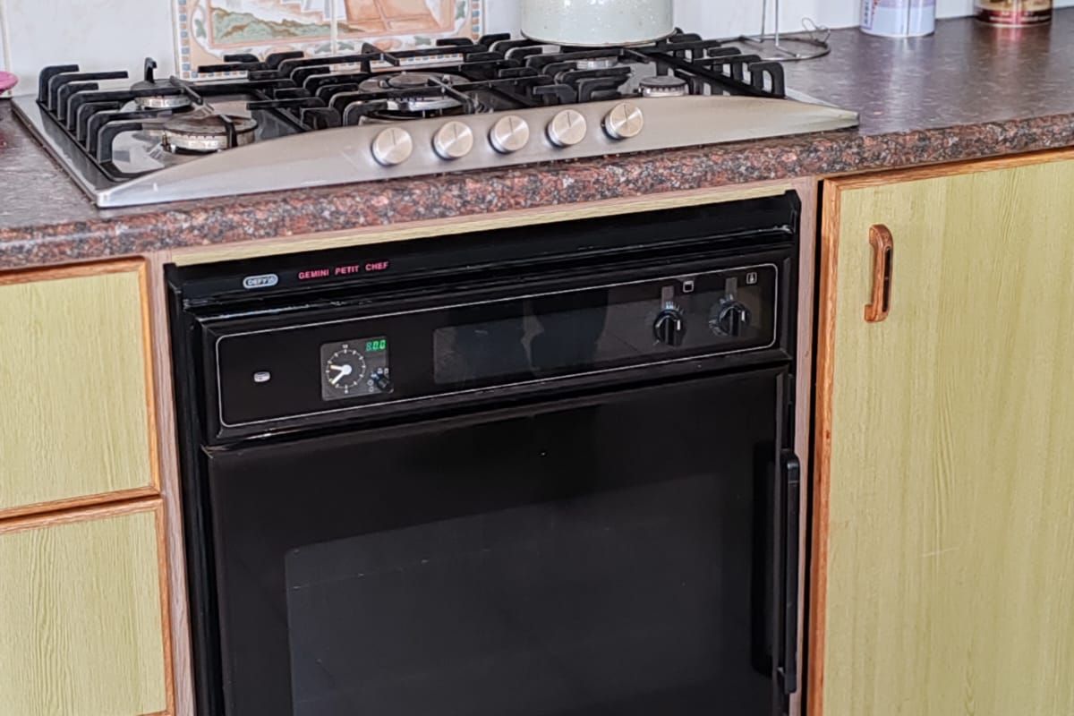 A close-up view of a kitchen workspace featuring a built-in oven, gas hob, tiled backsplash, and dated cabinetry.