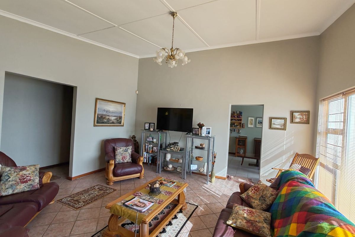 A spacious living room featuring tiled flooring, a central chandelier, and natural light filtering through venetian blinds, offering a comfortable and functional living space.