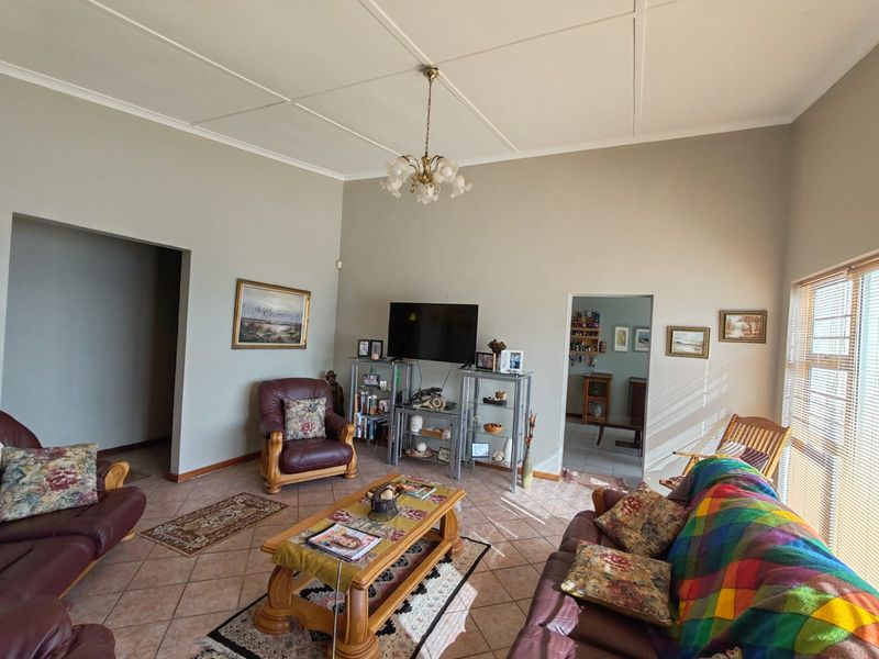 A spacious living room featuring tiled flooring, a central chandelier, and natural light filtering through venetian blinds, offering a comfortable and functional living space.