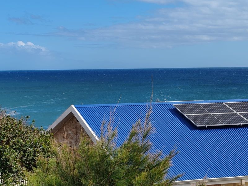 Elevated view of a blue corrugated metal roof featuring integrated solar panels, set against a scenic ocean backdrop.