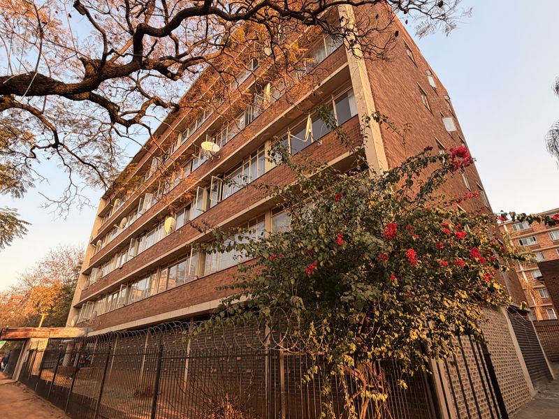 Exterior view of a multi-story brick apartment building featuring metal security fencing and mature trees in the foreground.