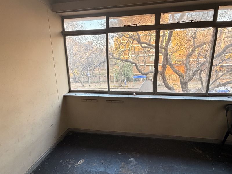 An empty, enclosed balcony area featuring large metal-framed windows with views of the surrounding neighborhood, requiring cosmetic updates to the walls and flooring.