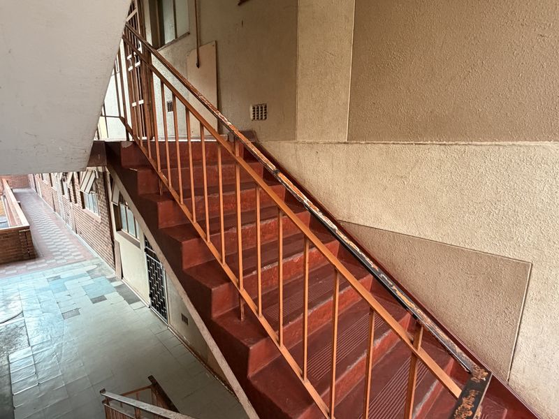 An open-air apartment building stairwell featuring a metal staircase and tiled flooring, showing signs of wear and aging.