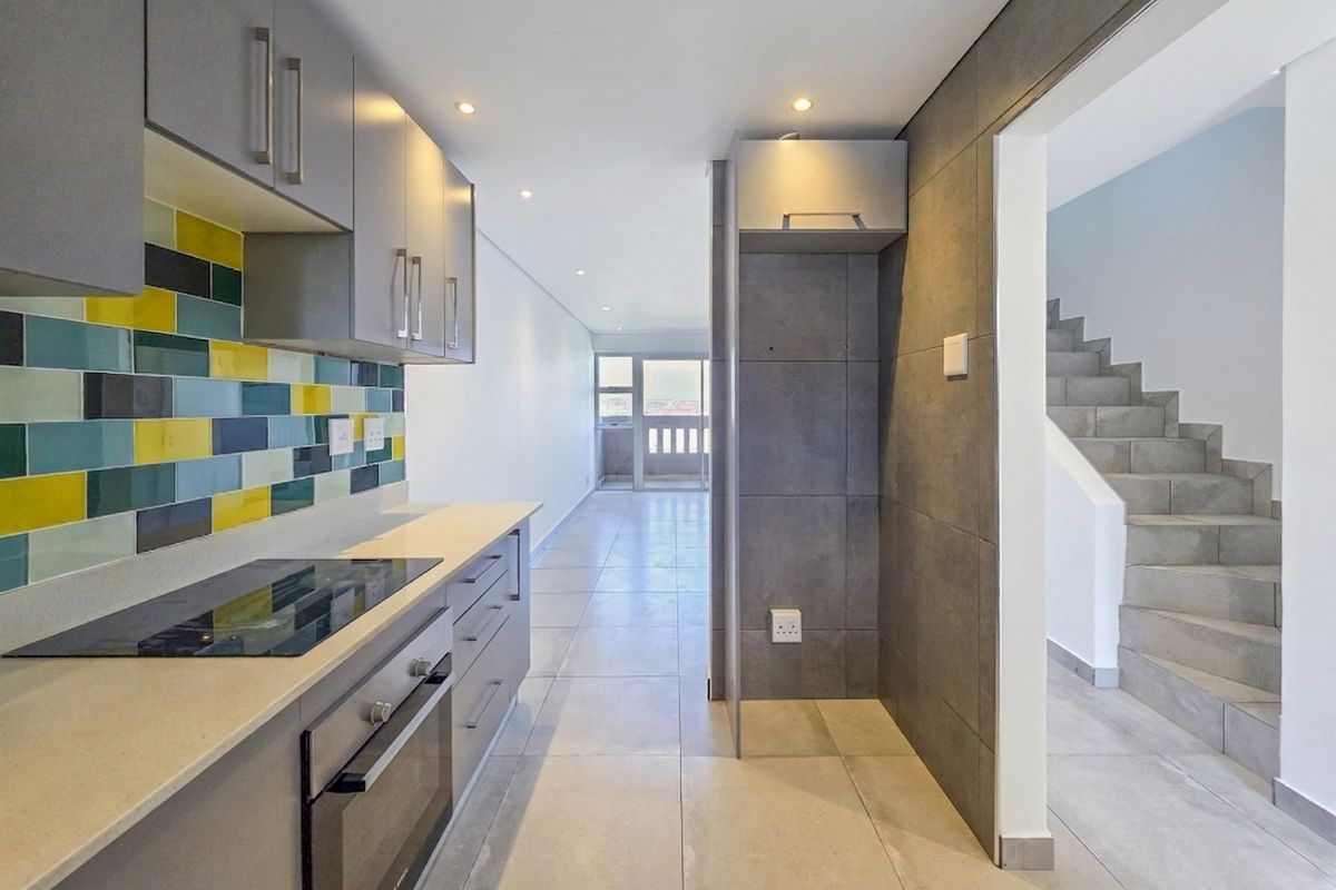 Modern kitchen featuring sleek grey cabinetry, stone countertops, integrated oven and hob, and a vibrant tiled splashback, leading to an open-plan living area.