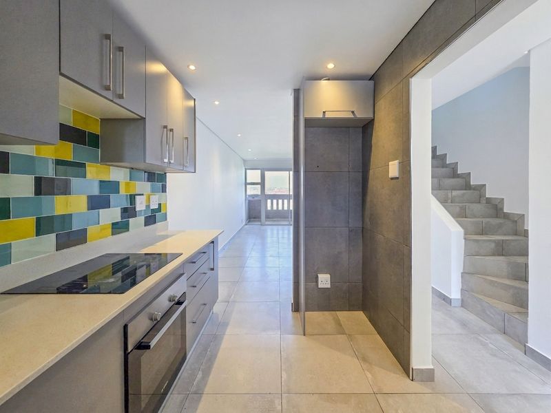 Modern kitchen featuring sleek grey cabinetry, stone countertops, integrated oven and hob, and a vibrant tiled splashback, leading to an open-plan living area.