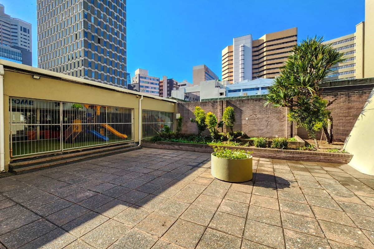 A paved outdoor courtyard area featuring a raised garden bed, brick boundary walls, and security fencing, set against an urban city backdrop.