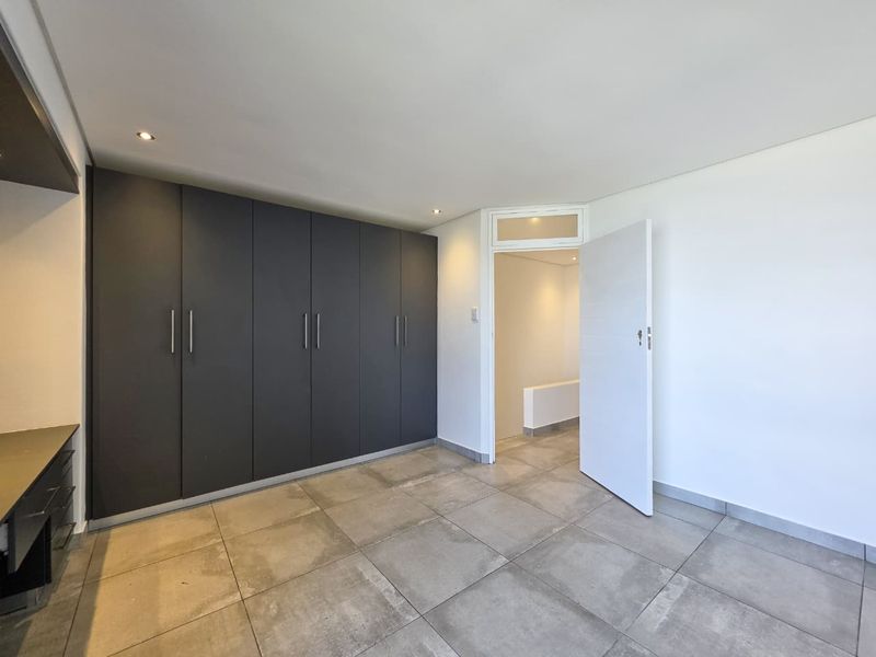 A bright, empty bedroom featuring modern dark grey built-in cupboards, large format floor tiles, and recessed ceiling lighting.