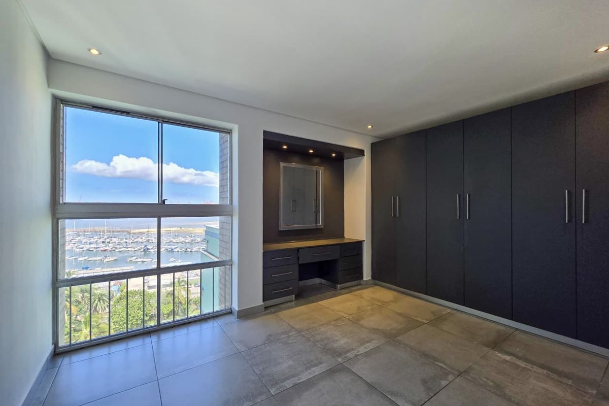 Spacious bedroom featuring dark built-in cupboards, a matching vanity unit, large windows with marina views, and modern tiled flooring.