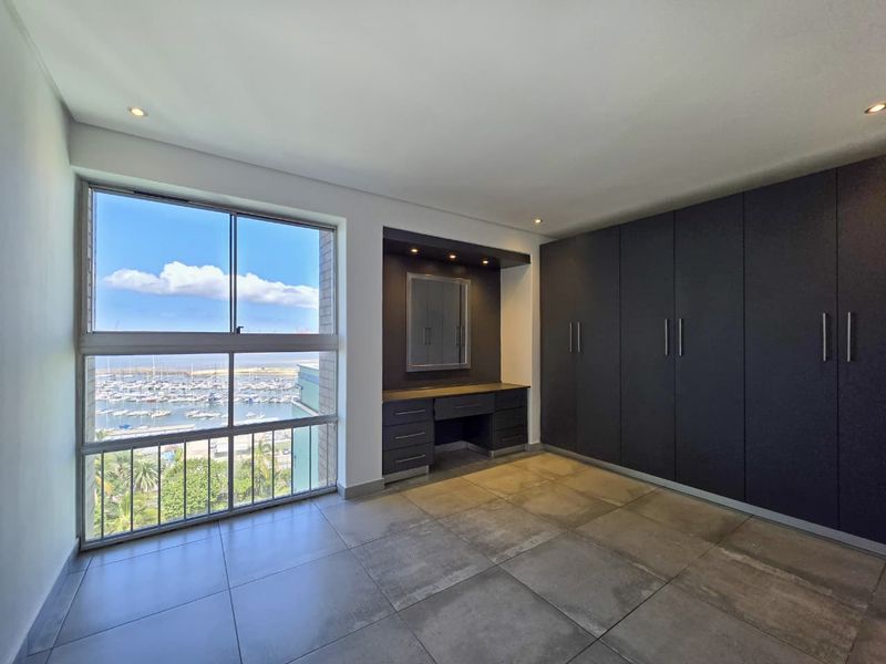 Spacious bedroom featuring dark built-in cupboards, a matching vanity unit, large windows with marina views, and modern tiled flooring.