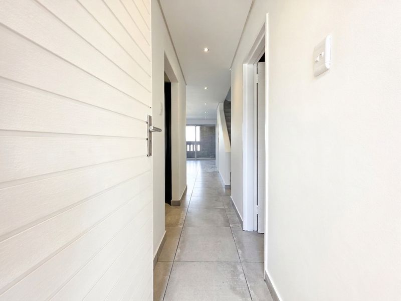 A bright, modern hallway featuring neutral tiled flooring, recessed downlighting, and clean white finishes leading towards the main living area.