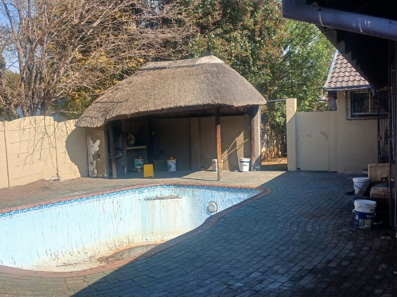 Outdoor entertainment area featuring a swimming pool, paved patio, and a thatch lapa with a built-in braai, requiring maintenance and refurbishment.