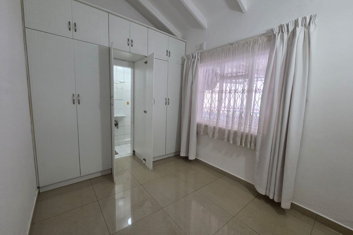 A bright, empty bedroom featuring tiled flooring, exposed ceiling beams, and extensive built-in cupboard storage with an integrated en-suite access.