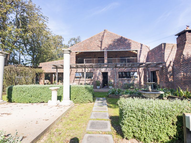 Rear view of a rustic brick house featuring a balcony, timber pergola, stone fountain, and established garden with a paved walkway.