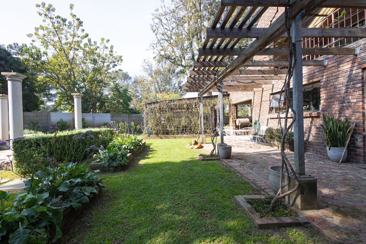 A spacious rear garden featuring a brick-paved patio with a timber pergola, raised garden beds, and established landscaping against a classic brick house facade.