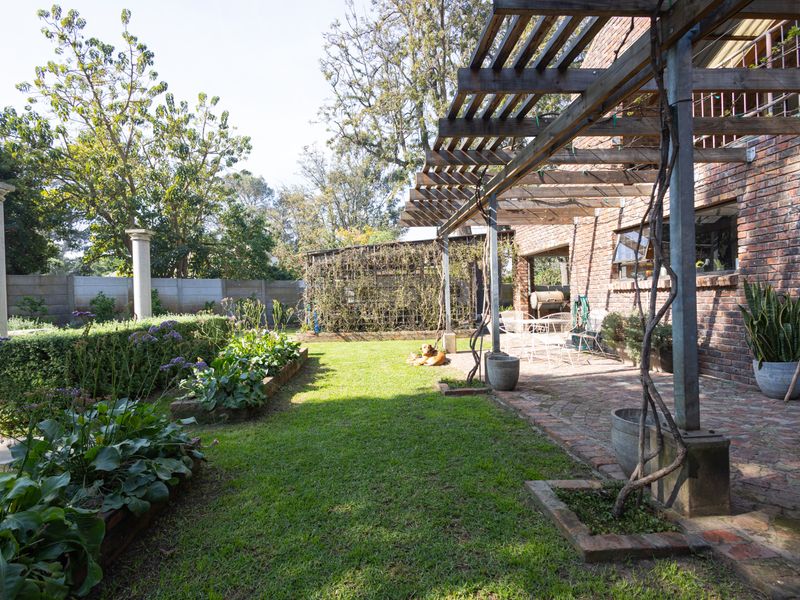 A spacious rear garden featuring a brick-paved patio with a timber pergola, raised garden beds, and established landscaping against a classic brick house facade.