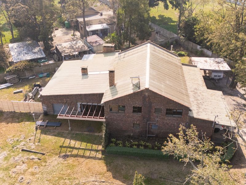Aerial view of a brick house featuring a corrugated metal roof, multiple chimneys, and an attached pergola structure set on a large plot.
