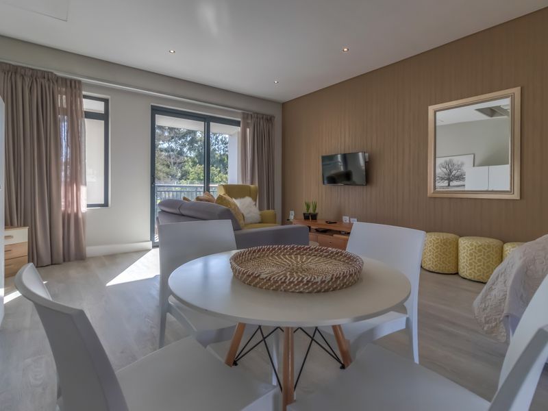 A bright open-plan living and bedroom area featuring modern laminate flooring, recessed lighting, and direct access to a private balcony through sliding glass doors.