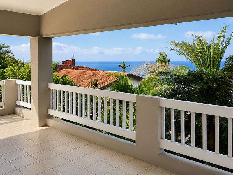 A spacious covered balcony featuring tiled flooring and a white balustrade, offering elevated views of the surrounding greenery and the ocean.