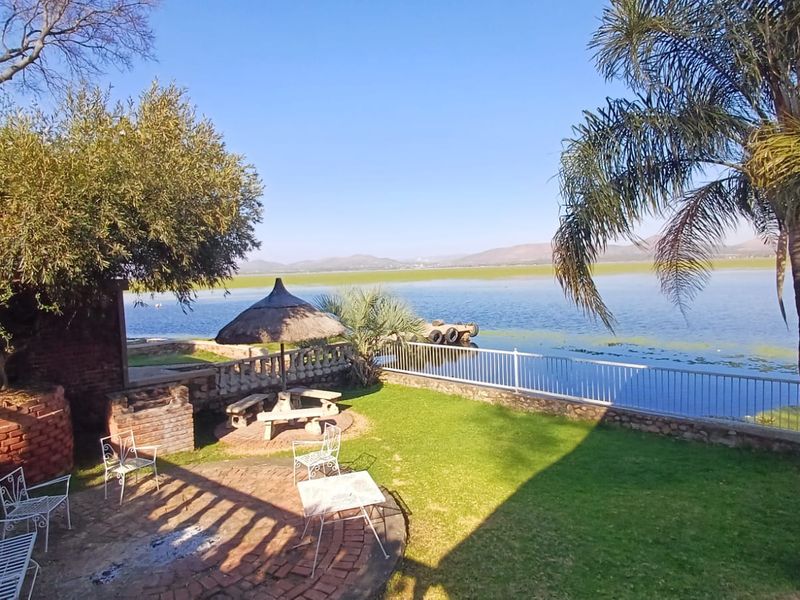 A scenic outdoor garden area featuring a brick braai, stone paving, and a metal balustrade overlooking a large lake with mountain views.