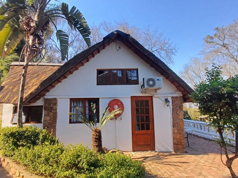 A charming cottage-style exterior featuring a traditional thatch roof, stone accents, and a paved patio area, set within a landscaped garden.