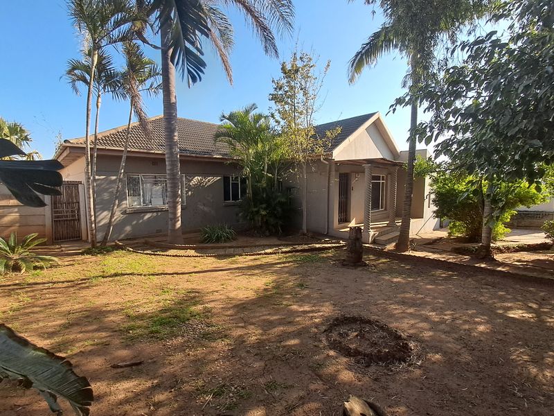 Front view of a single-story residential house featuring a tiled roof, covered entrance porch, and established garden with mature palm trees.