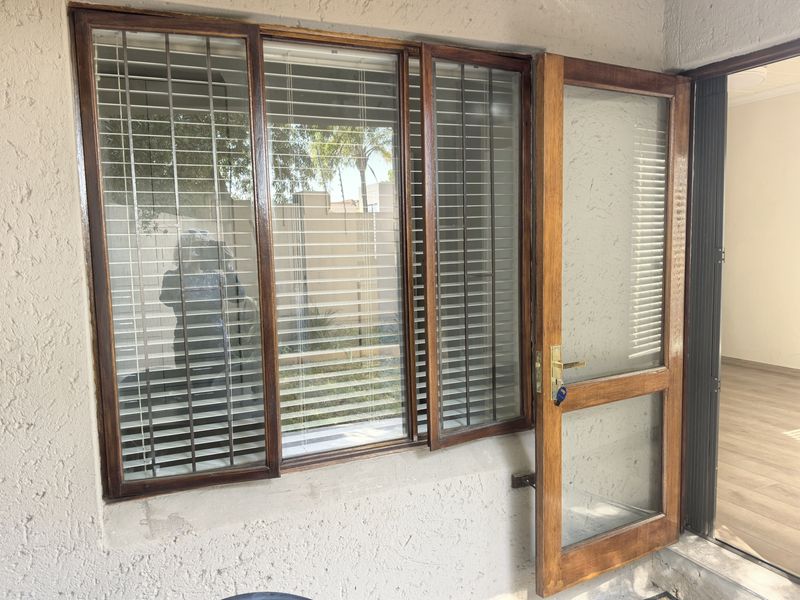 A covered patio area featuring timber-framed windows and a glass door, showing signs of wear on the wood finishes and exterior wall.