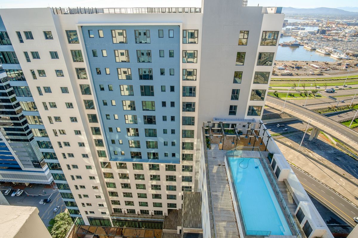 Aerial view of a modern high-rise apartment building featuring a rooftop swimming pool, timber decking, and glass balustrades overlooking the harbor.