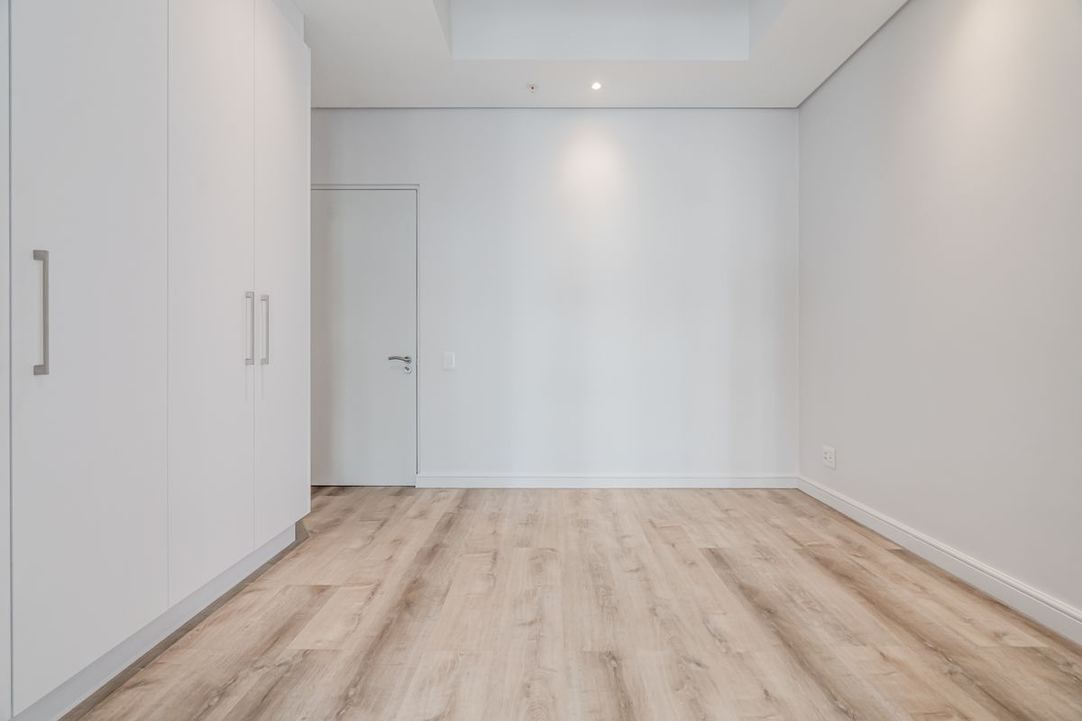A bright, empty bedroom featuring modern laminate flooring, built-in cupboards, and a clean, minimalist aesthetic with recessed ceiling lighting.