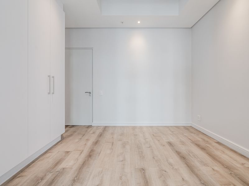 A bright, empty bedroom featuring modern laminate flooring, built-in cupboards, and a clean, minimalist aesthetic with recessed ceiling lighting.
