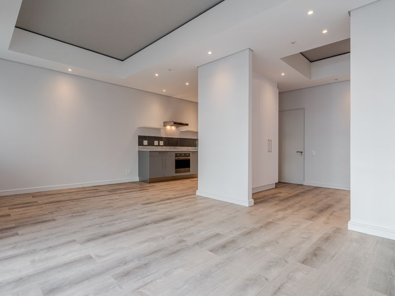 Modern open-plan living area featuring light laminate flooring, recessed ceiling details with downlights, and a compact kitchen with built-in cabinetry and appliances.
