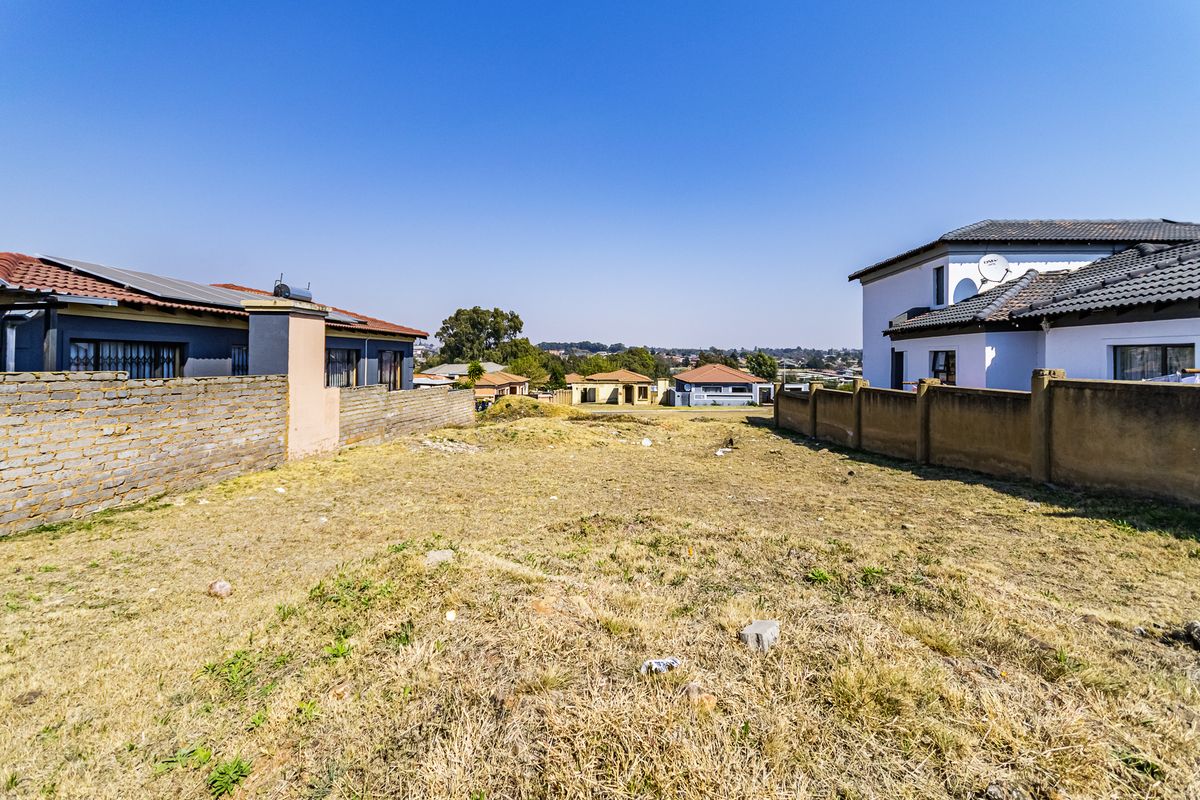 A vacant residential plot situated between existing neighboring properties, featuring dry ground and perimeter boundary walls.