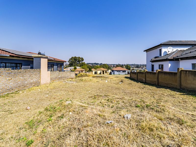 A vacant residential plot situated between existing neighboring properties, featuring dry ground and perimeter boundary walls.