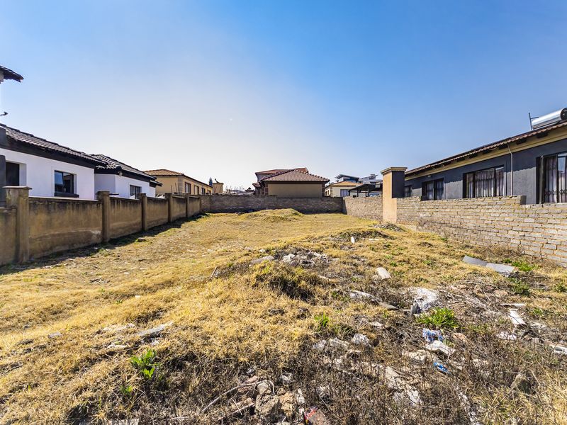 A vacant residential plot enclosed by perimeter brick boundary walls, featuring uneven terrain with dry grass and scattered debris.